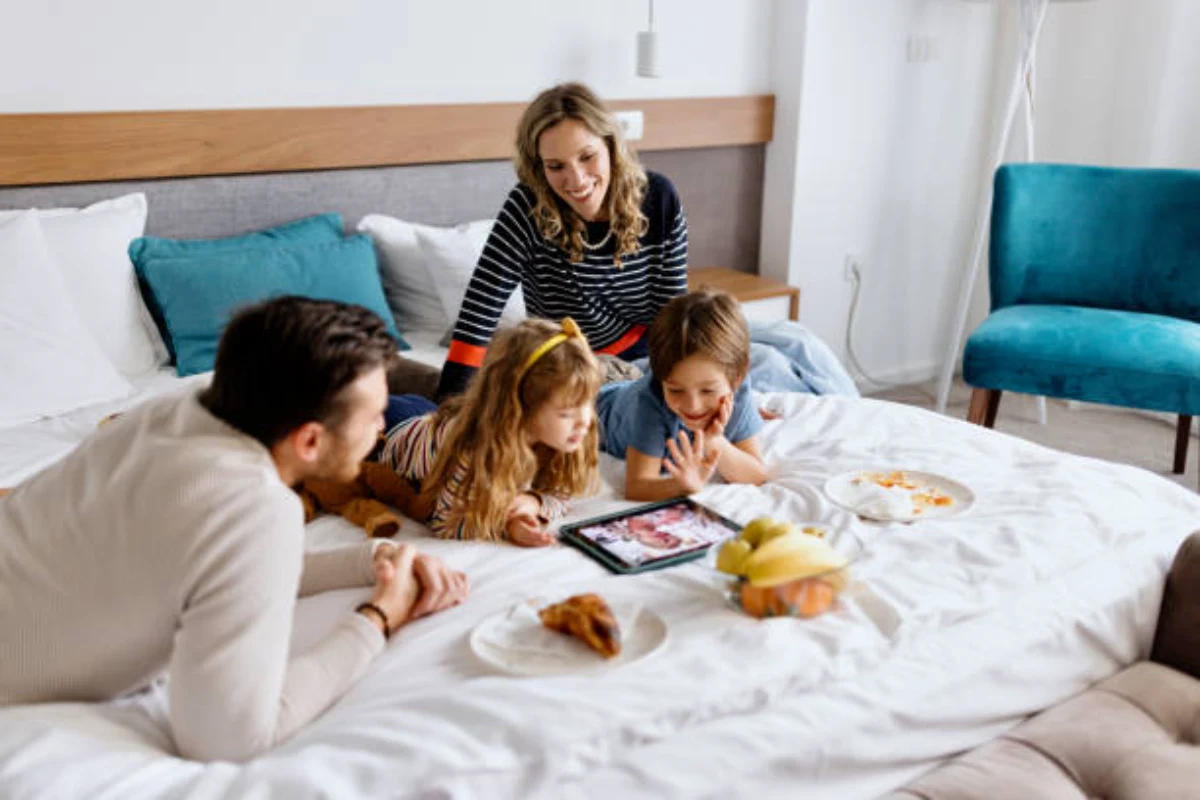 Family enjoying breakfast and screen time together on a hotel bed during a staycation.