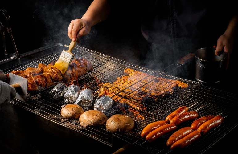 Chef grilling BBQ meats, sausages, mushrooms, and baked potatoes on a smoky rooftop grill.